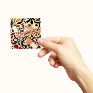 Hånd som holder et firkantet klistremerke med blomster og teksten never stop dreaming